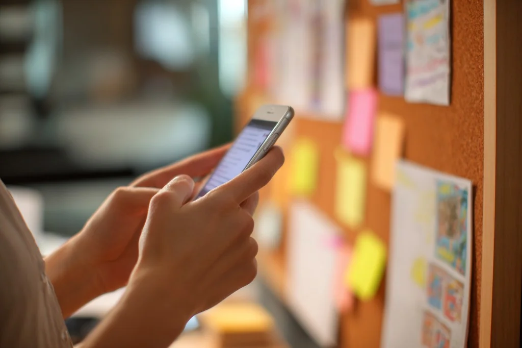 Hands on a smartphone beside a planning corkboard, suggesting private coordination without sharing contact lists